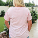 Woman with blonde hair standing in a back profile position wearing the Basic Tee in a lifestyle scene. The focal point of this image is the pink tee and length of tee. The model paired this tee with a light-wash denim skirt. 