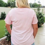 Woman with blonde hair standing in a back profile position wearing the Basic Tee in a lifestyle scene. The focal point of this image is the pink tee and length of tee. The model paired this tee with a light-wash denim skirt. 