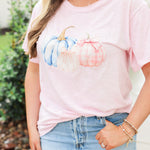 Woman with blonde hair standing in a front profile position wearing the Basic Tee in a lifestyle scene. The focal point of this image is the pink and blue pumpkin graphics. The model paired this top with a light-wash denim skirt. 