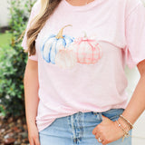 Woman with blonde hair standing in a front profile position wearing the Basic Tee in a lifestyle scene. The focal point of this image is the pink and blue pumpkin graphics. The model paired this top with a light-wash denim skirt. 