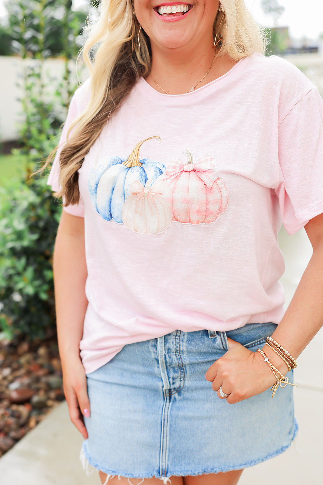 Woman with blonde hair standing in a front profile position wearing the Basic Tee in a lifestyle scene. The focal point of this image is the pink and blue pumpkin graphics. The model paired this top with a light-wash denim skirt. 