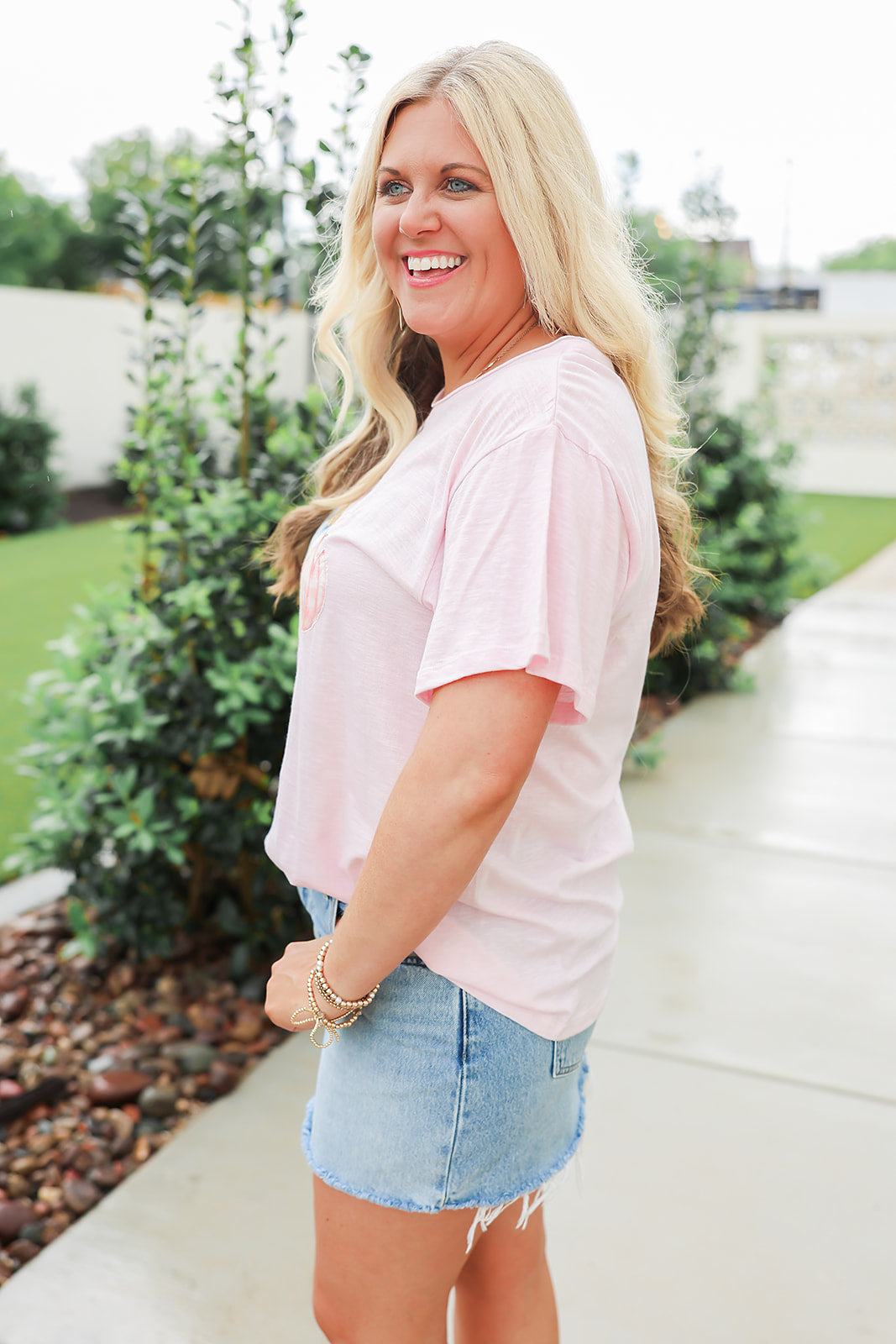 Woman with blonde hair standing in a side profile position wearing the Basic Tee in a lifestyle scene. The focal point of this image is the side profile of tee. The model paired this tee with light-wash denim jeans. 