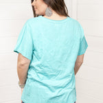 Woman with dark hair standing in a back profile position wearing the Basic Tee against a white background. The focal point of this image is the seaside blue tee. The model paired this tee with dark-wash denim shorts. 