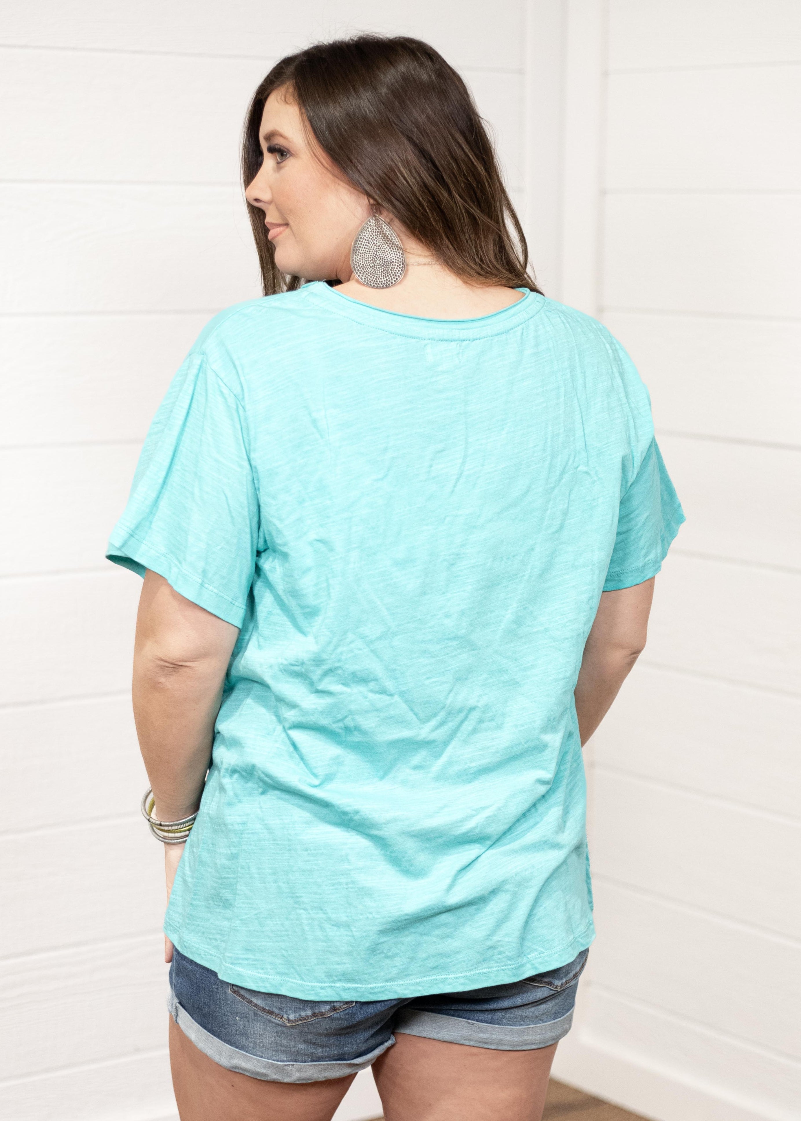 Woman with dark hair standing in a back profile position wearing the Basic Tee against a white background. The focal point of this image is the seaside blue tee. The model paired this tee with dark-wash denim shorts. 