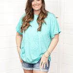 Woman with dark hair standing in front profile position wearing the Basic Tee against a white background. The focal point of this image is the seaside blue colored tee. The model paired this tee with dark-wash denim shorts. 