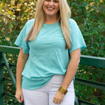 Woman with blonde hair standing in front profile position wearing the Basic Tee in a lifestyle scene. The focal point of this image is the seaside blue colored tee. The model paired this tee with white denim pants. 