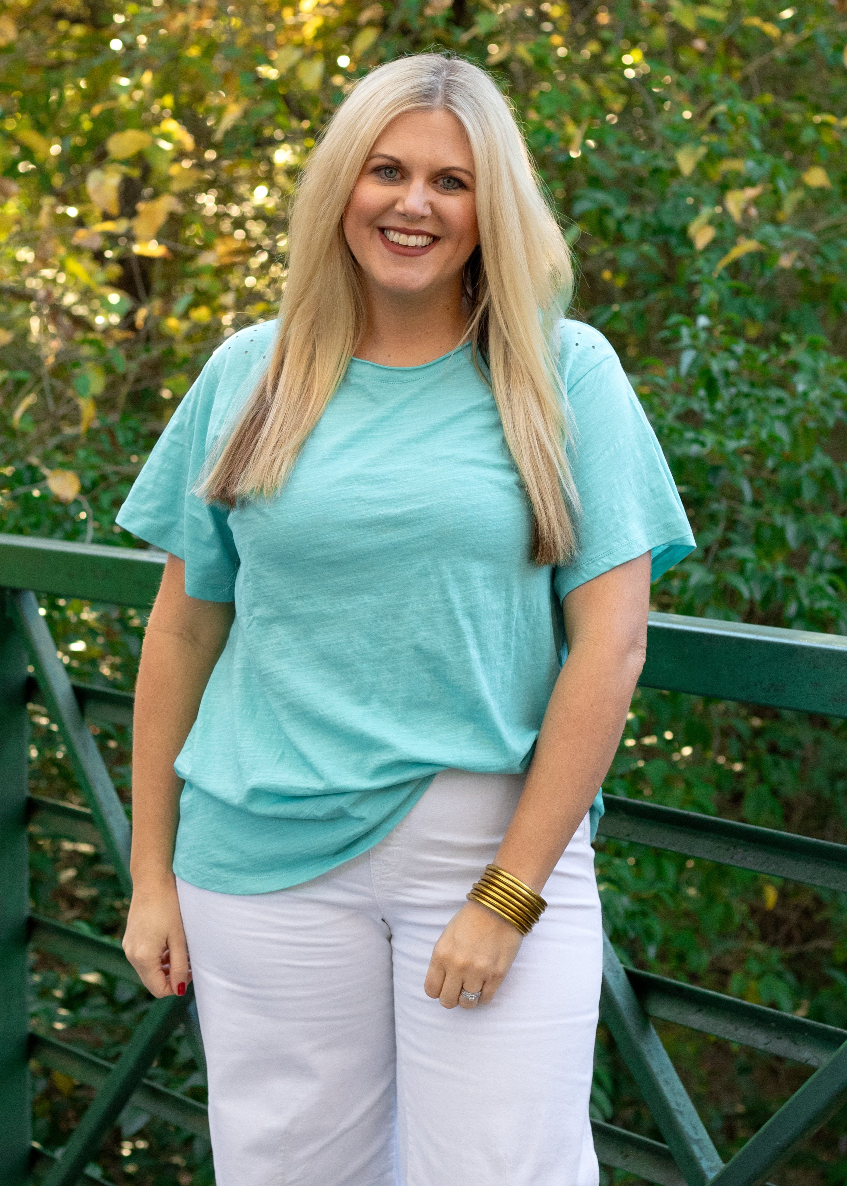 Woman with blonde hair standing in front profile position wearing the Basic Tee in a lifestyle scene. The focal point of this image is the seaside blue colored tee. The model paired this tee with white denim pants. 