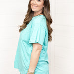 Woman with dark hair standing in a side profile position wearing the Basic Tee against a white background. The focal point of this image is the seaside blue colored tee. The model paired this tee with dark-wash denim shorts.