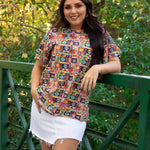 Woman with dark hair standing in a front profile position wearing the Basic Tee in a lifestyle scene. The focal point of this image is the graphics on tee in a heavy greenery setting. The model paired this top with a white denim skirt. 