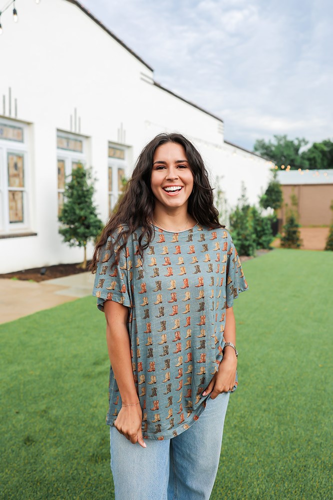 Woman with dark hair standing in a front profile position wearing the Basic Tee in a lifestyle scene. The focal point of this image is the cowboy boot graphics on front of tee. The model paired this top with light-wash denim jeans. 