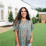 Woman with dark hair standing in a front profile position wearing the Basic Tee in a lifestyle scene. The focal point of this image is the cowboy boot graphics on front of tee. The model paired this top with light-wash denim jeans. 