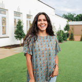 Woman with dark hair standing in a front profile position wearing the Basic Tee in a lifestyle scene. The focal point of this image is the cowboy boot graphics on front of tee. The model paired this top with light-wash denim jeans. 