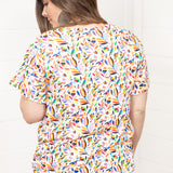 Woman with dark hair standing in a back profile position wearing the Basic Tee against a white background. The focal point of this image is colorful otomi graphics. The model paired this top with dark-wash denim shorts. 