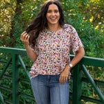 Woman with dark hair standing in a front profile position wearing the Basic Tee in a lifestyle scene. The focal point of this image is the colorful otomi graphics against heavy greenery. The model paired this top with dark-wash denim pants.