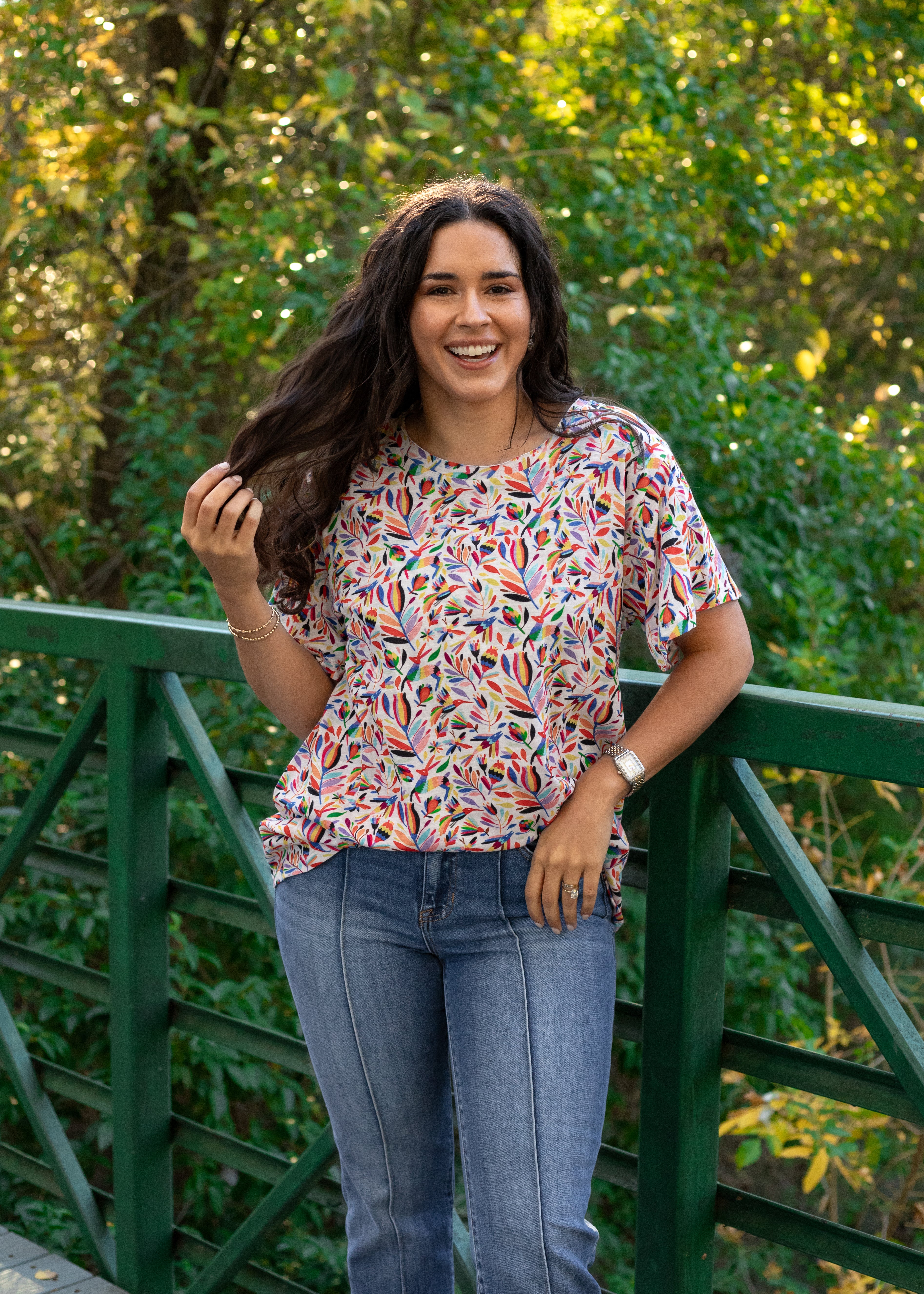 Woman with dark hair standing in a front profile position wearing the Basic Tee in a lifestyle scene. The focal point of this image is the colorful otomi graphics against heavy greenery. The model paired this top with dark-wash denim pants.