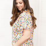 Woman with dark hair standing in a side profile position wearing the Basic Tee against a white background. The focal point of this image is the color otomi pattern. The model paired this top with dark-wash denim shorts. 