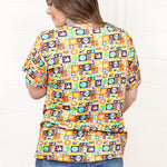 Woman with dark hair standing in a back profile position wearing the Basic Tee against a white background. The focal point of this image is the bright-colored graphics that include sugar skulls, cacti, etc. The model paired this tee with dark-wash denim shorts.