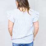 Woman with dark hair standing in a back profile position wear the Beck Top against a white background. The focal point of this image is the blue striped graphic of the shirt, ruffles sleeves, and red embroidery along neckline. The model paired this top with dark-wash denim shorts.