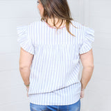 Woman with dark hair standing in a back profile position wear the Beck Top against a white background. The focal point of this image is the blue striped graphic of the shirt, ruffles sleeves, and red embroidery along neckline. The model paired this top with dark-wash denim shorts.