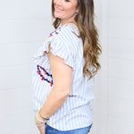 Woman with dark hair standing in a side profile position wearing the Beck Top against a white background. The focal point of this image is the ruffled sleeves, blue striped fabric and red and navy embroidery slightly seen on chest of top. The model paired this top with dark-wash denim shorts.