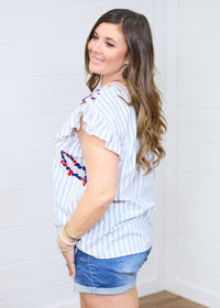 Woman with dark hair standing in a side profile position wearing the Beck Top against a white background. The focal point of this image is the ruffled sleeves, blue striped fabric and red and navy embroidery slightly seen on chest of top. The model paired this top with dark-wash denim shorts.