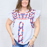 Woman with dark hair standing in a front profile position wearing the Beck Top against a white background. The focal point of this image is the red and blue floral embroidery on front of top and red sticking along neckline. The model paired the top with dark-wash denim shorts.