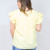 Woman with dark hair standing in a back profile position wearing the Beck Top against a white background. The focal point of this image is to show ruffles around sleeves and pleating below shoulders. The model paired this top with dark-wash denim shorts.