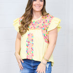 Woman with dark hair standing in a front profile position wearing the Beck Top against a white background. The focal point of this image is the embroidery along middle of the top and shoulders. The model paired this top with dark-wash denim shorts.