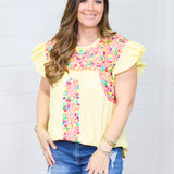 Woman with dark hair standing in a front profile position wearing the Beck Top against a white background. The focal point of this image is the embroidery along middle of the top and shoulders. The model paired this top with dark-wash denim shorts.