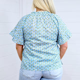 Woman with blonde hair standing in a back profile position wearing the Bell Top against a white background. The focal point of this image is the green and blue graphics, pleating on sleeves and top of collar. the model paired this top with light-wash denim shorts. 