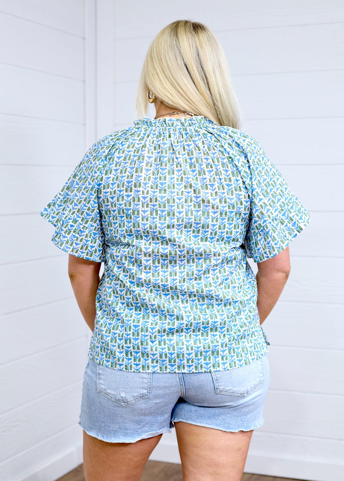 Woman with blonde hair standing in a back profile position wearing the Bell Top against a white background. The focal point of this image is the green and blue graphics, pleating on sleeves and top of collar. the model paired this top with light-wash denim shorts. 