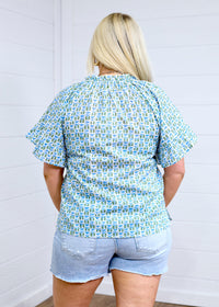 Woman with blonde hair standing in a back profile position wearing the Bell Top against a white background. The focal point of this image is the green and blue graphics, pleating on sleeves and top of collar. the model paired this top with light-wash denim shorts. 