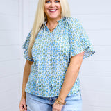 Woman with blonde hair standing in a front profile position wearing the Bell Top against a white background. The focal point of this image is the graphics on top as well as sleeve ruffles and collar ruffles. The model paired this top with light-wash denim shorts. 