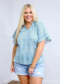 Woman with blonde hair standing in a front profile position wearing the Bell Top against a white background. The focal point of this image is the graphics on top as well as sleeve ruffles and collar ruffles. The model paired this top with light-wash denim shorts. 