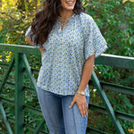 Woman with dark hair standing in a front profile position wearing the Bell Top in a lifestyle scene. The focal point of this image is the graphics , pleated collar, and tassels that tie in front. The model paired this top with dark-wash denim jeans. 