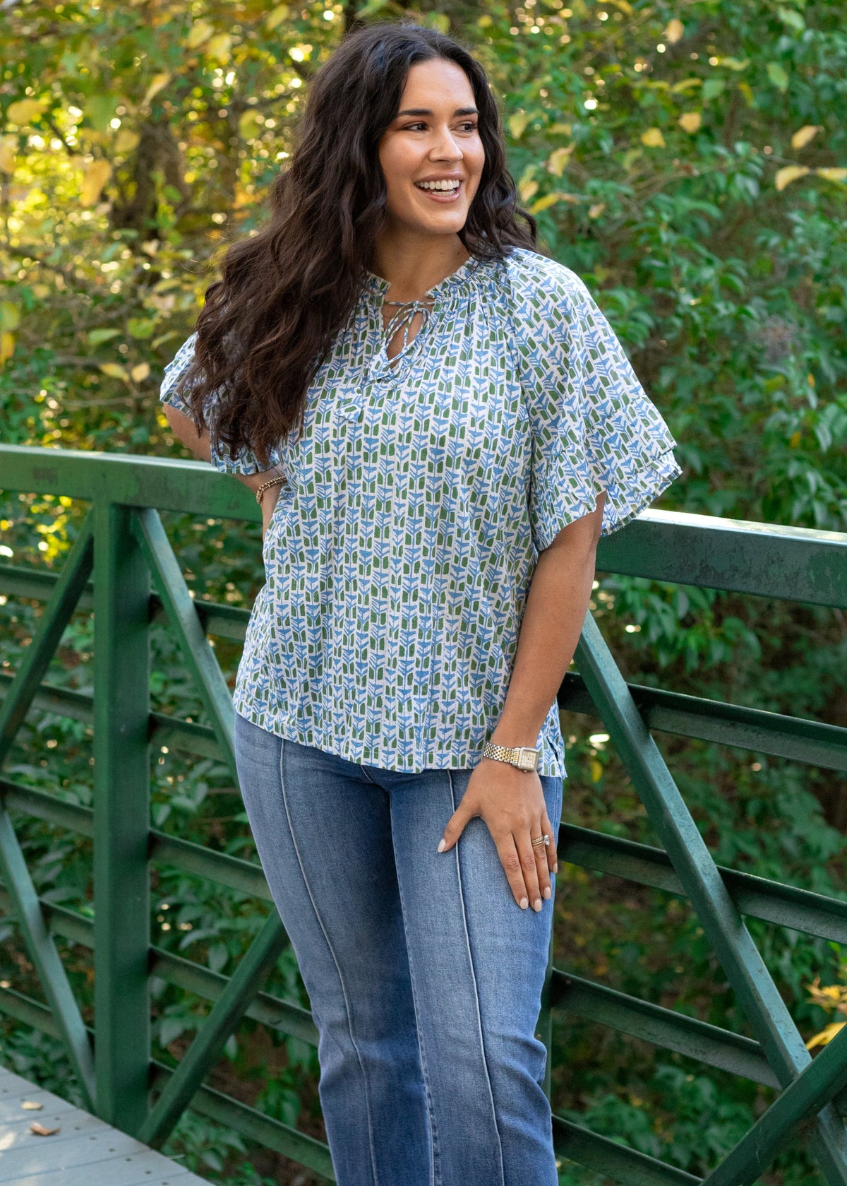 Woman with dark hair standing in a front profile position wearing the Bell Top in a lifestyle scene. The focal point of this image is the graphics , pleated collar, and tassels that tie in front. The model paired this top with dark-wash denim jeans. 