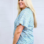 Woman with blonde hair standing in a side profile position wearing the Bell Top against a white background. The focal point of this image is the blue and green graphics, sleeve length. The model paired this top with light-wash denim shorts. 
