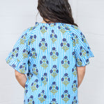 Woman with dark hair standing in a back profile position wearing the Bell Top against a white background. The focal point of this image is the graphics on the back, emphasizing the blue flowers and stripes. The model paired this  top with dark-wash denim jeans.