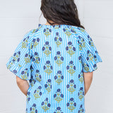 Woman with dark hair standing in a back profile position wearing the Bell Top against a white background. The focal point of this image is the graphics on the back, emphasizing the blue flowers and stripes. The model paired this  top with dark-wash denim jeans.