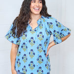 Woman with dark hair standing in a front profile position wearing the Bell Top against a white background. The focal point of the image is the graphics and rouse on collar of top. The model paired this top with dark-wash denim jeans. 