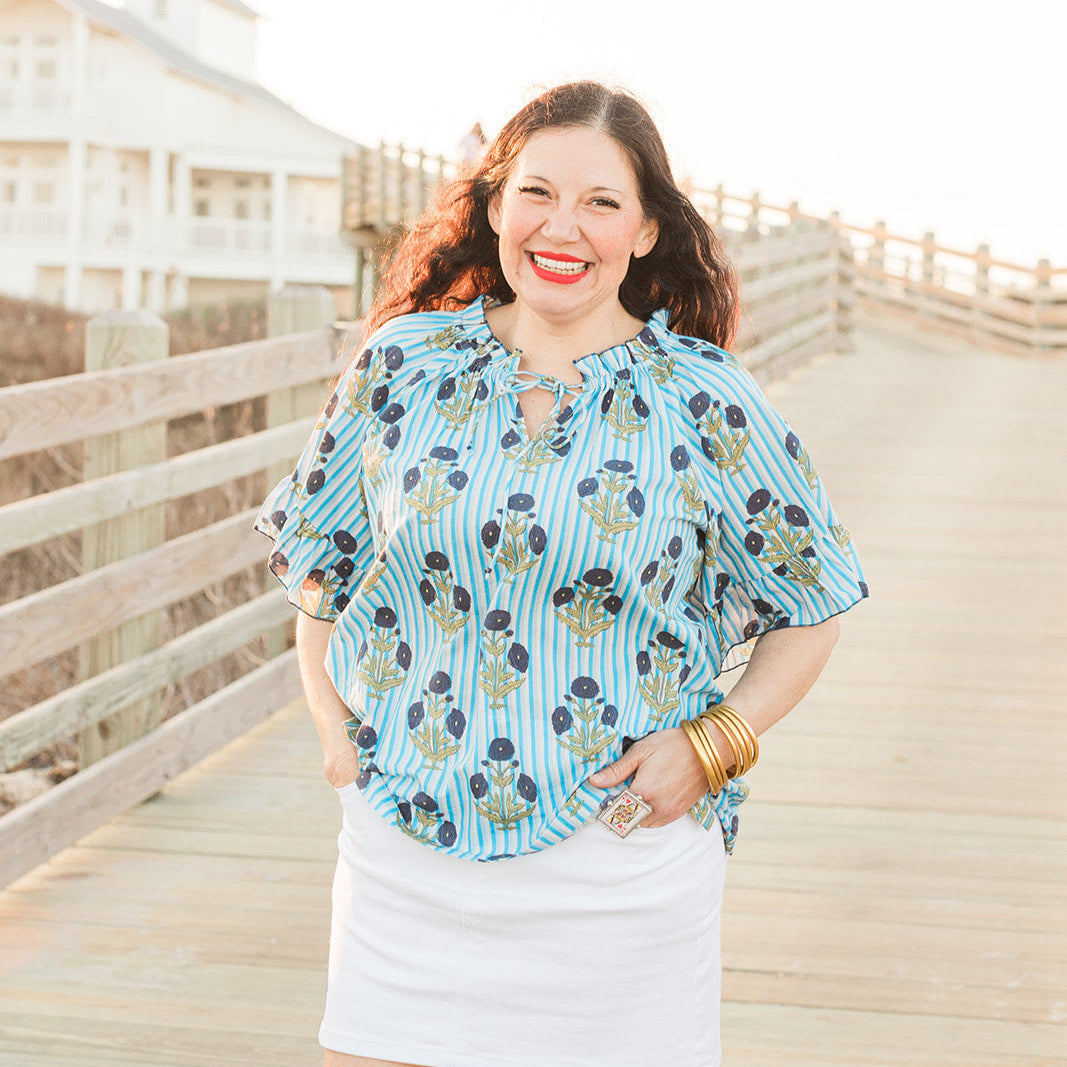 Woman with red hair standing in a front profile position wearing the Bell Top in a lifestyle scene. The focal point of this image is to see the top in its entirety. The model paired this top with a white skirt. 