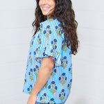 Woman with dark hair standing in a side profile position wearing the Bell Top against a white background. The focal point of this image is the graphics down the side of the top and the ruffle on the sleeve. The model paired this top with dark-wash denim jeans.