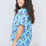 Woman with dark hair standing in a side profile position wearing the Bell Top against a white background. The focal point of this image is the graphics down the side of the top and the ruffle on the sleeve. The model paired this top with dark-wash denim jeans.