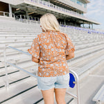 Woman with blonde hair standing in a back profile position wearing the Bell Top at a football field. The focal point of this image is the floral graphics on back and shoulders as well as the ruffled sleeves. The model paired this top with light-wash denim shorts.