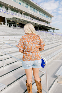 Woman with blonde hair standing in a back profile position wearing the Bell Top at a football field. The focal point of this image is the floral graphics on back and shoulders as well as the ruffled sleeves. The model paired this top with light-wash denim shorts.