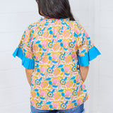 Woman with dark hair standing in a back profile position wearing the Bell Top against a white background. The focal point of this image is the pink, blue, orange, and green graphics, as well as blue ruffled sleeves. The model paired this top with dark-wash denim shorts.