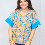 Woman with dark hair standing in a front profile position wearing the Bell Top against a white background. The focal point for this image is the graphics on front of top, ruffled sleeves, v-neckline and tassels. The model paired this top with dark-wash denim shorts.