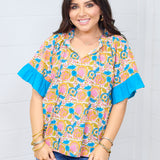 Woman with dark hair standing in a front profile position wearing the Bell Top against a white background. The focal point for this image is the graphics on front of top, ruffled sleeves, v-neckline and tassels. The model paired this top with dark-wash denim shorts.