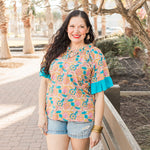 Woman with dark hair standing in a front profile position wearing the Bell Top in a lifestyle scene. The focal point of this image is to see the top in its entirety. The model paired this top with light-wash denim shorts.