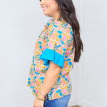 Woman with dark hair standing in side profile position wearing the Bell Top against a white background. The focal point of this top is to see the length of the top, side graphics, and ruffles on sleeve. The model paired this top with dark-wash denim shorts.